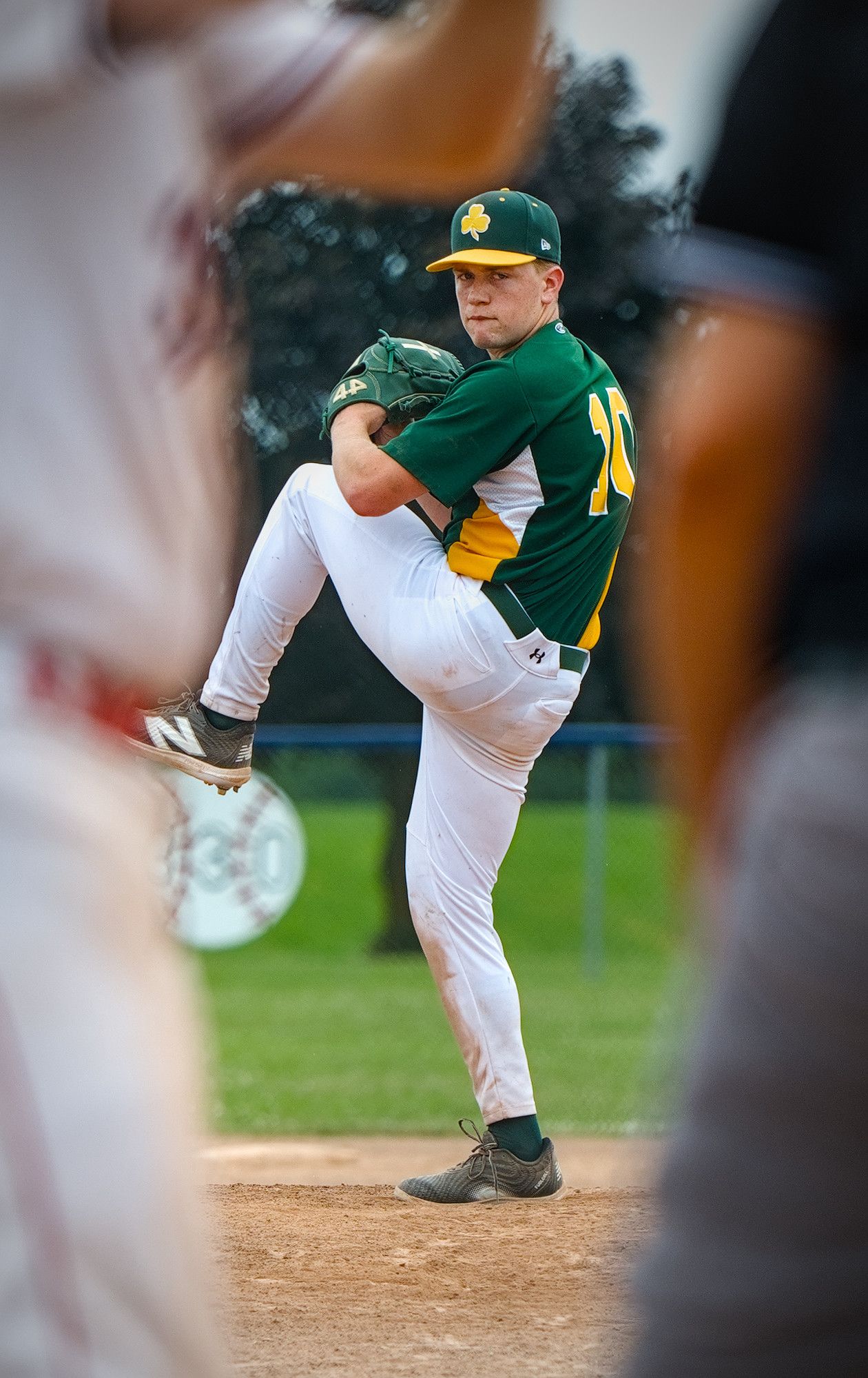 A pitcher at the top of his leg kick, glove tucked to his chest and eyes locked on the plate, framed between two out-of-focus figures in the foreground.