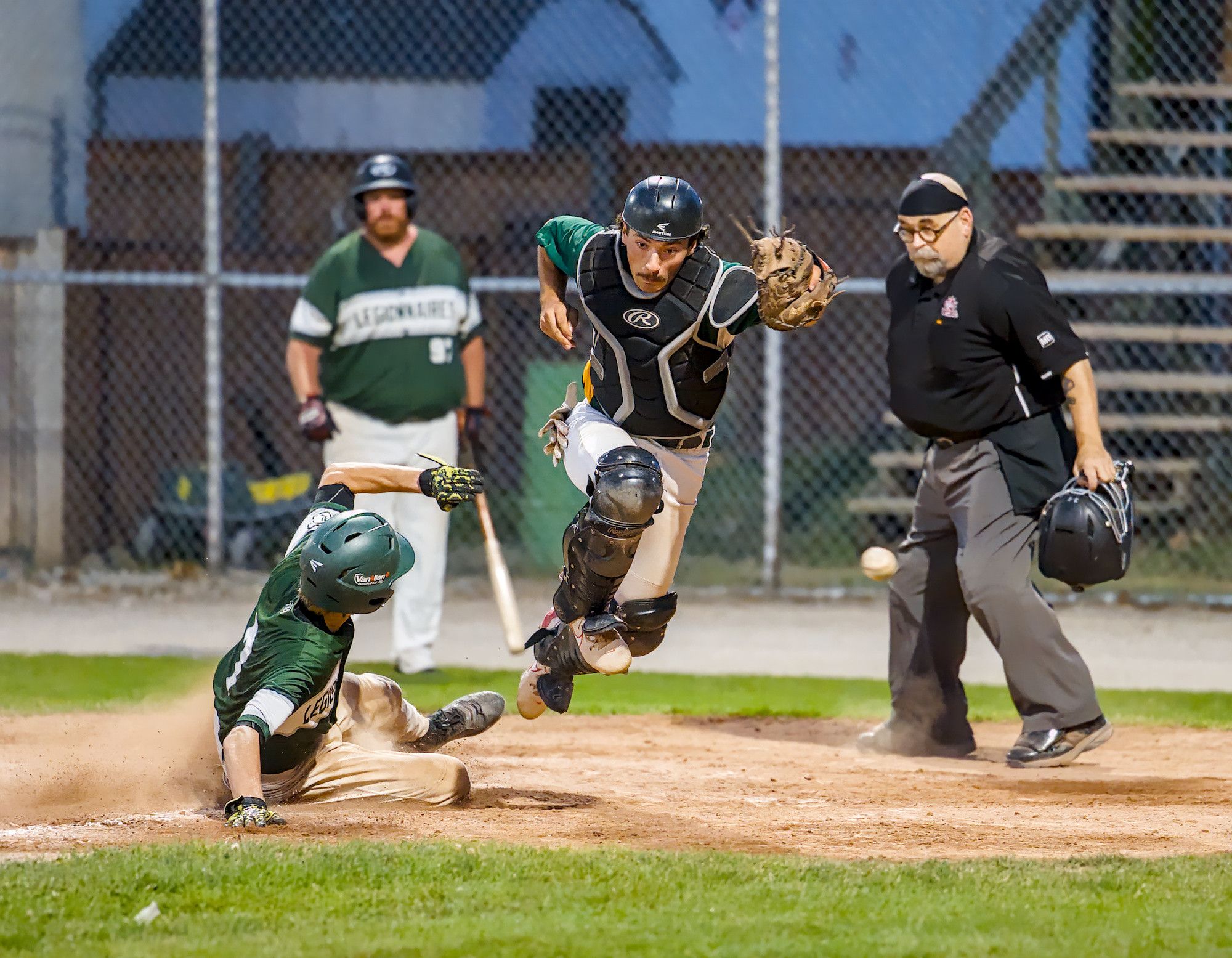A catcher leaps over a runner sliding into home plate as the ball hangs in mid-air beside the umpire at dusk.