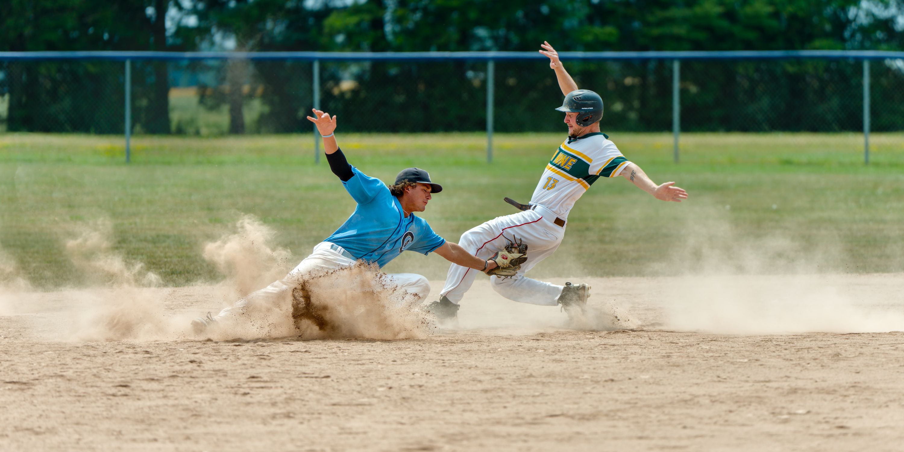 A fielder in blue lunges to tag a runner sliding into second base through an exploding cloud of infield dust, the runner's arms thrown wide.