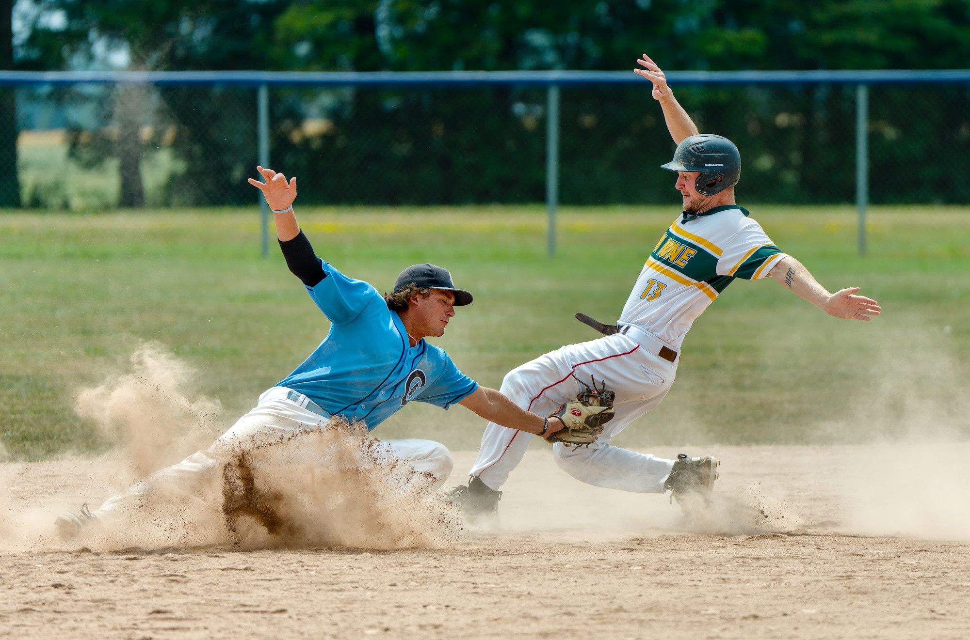 A fielder in blue lunges to tag a runner sliding into second base through an exploding cloud of infield dust, the runner's arms thrown wide.