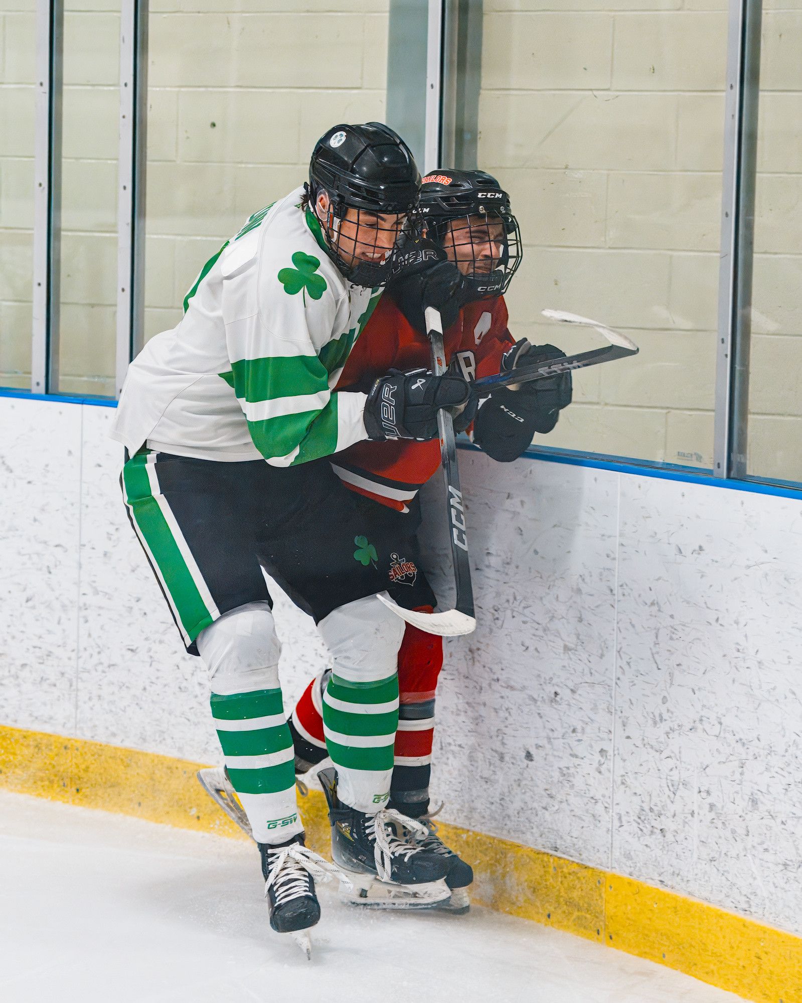 A player in a white shamrock jersey pins an opponent in red against the boards, both sticks tangled between them.