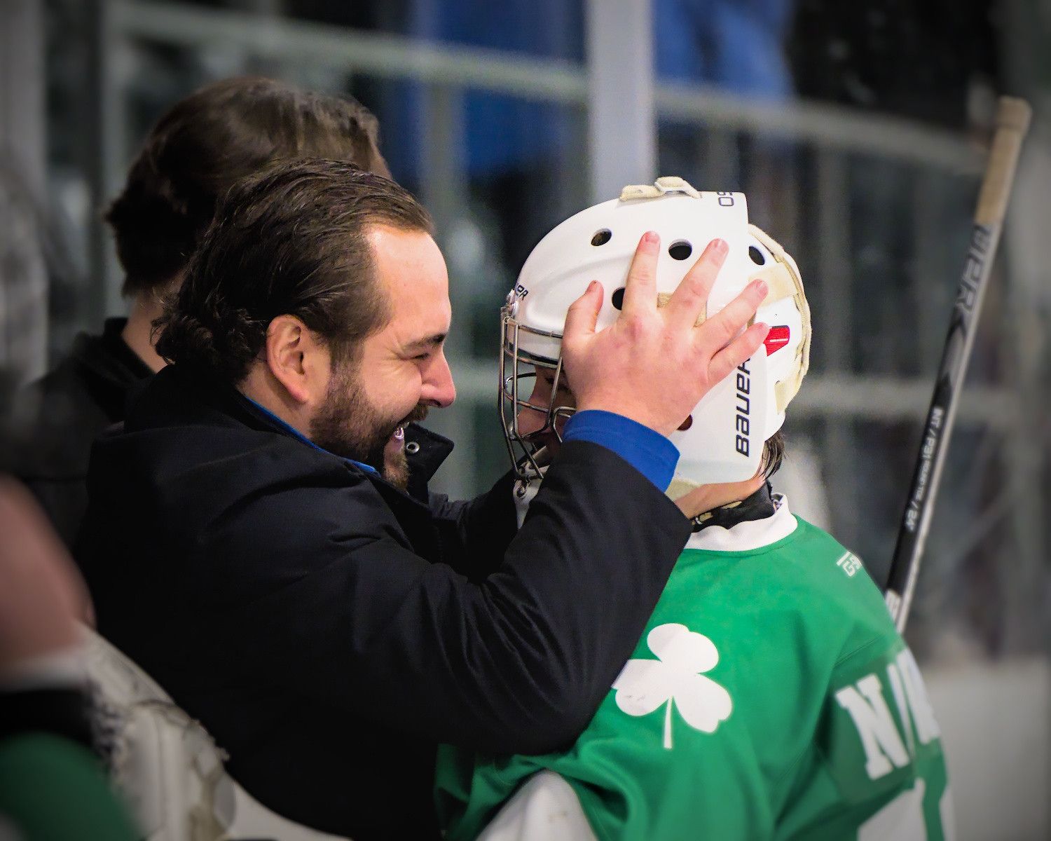 A smiling coach reaches across and grips his goaltender's mask with one hand in a bench celebration.