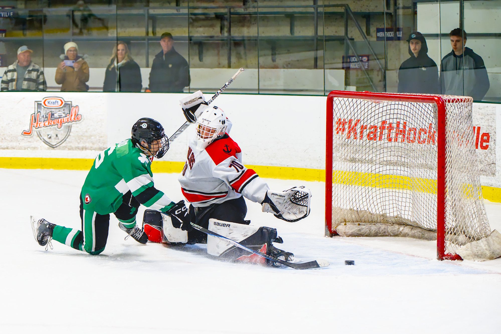 A goaltender sprawls across the crease with a pad stretched out while an attacker slides in, the puck loose on the ice by the post.