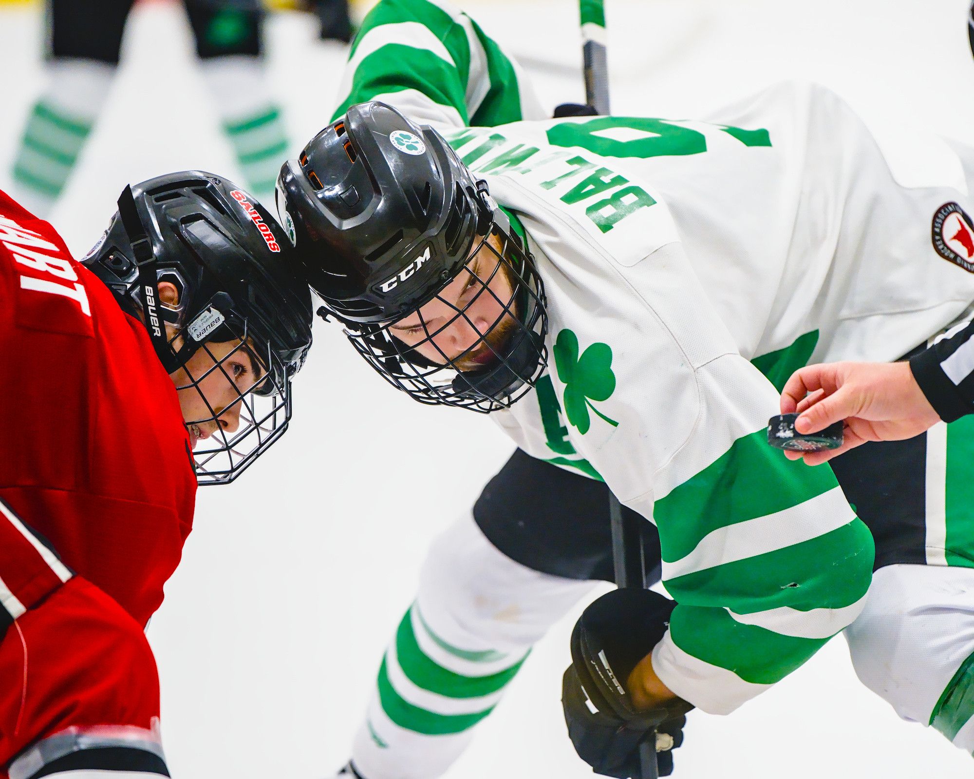 Two players lean helmet to helmet over the faceoff dot as the linesman's hand holds the puck at the edge of the frame.