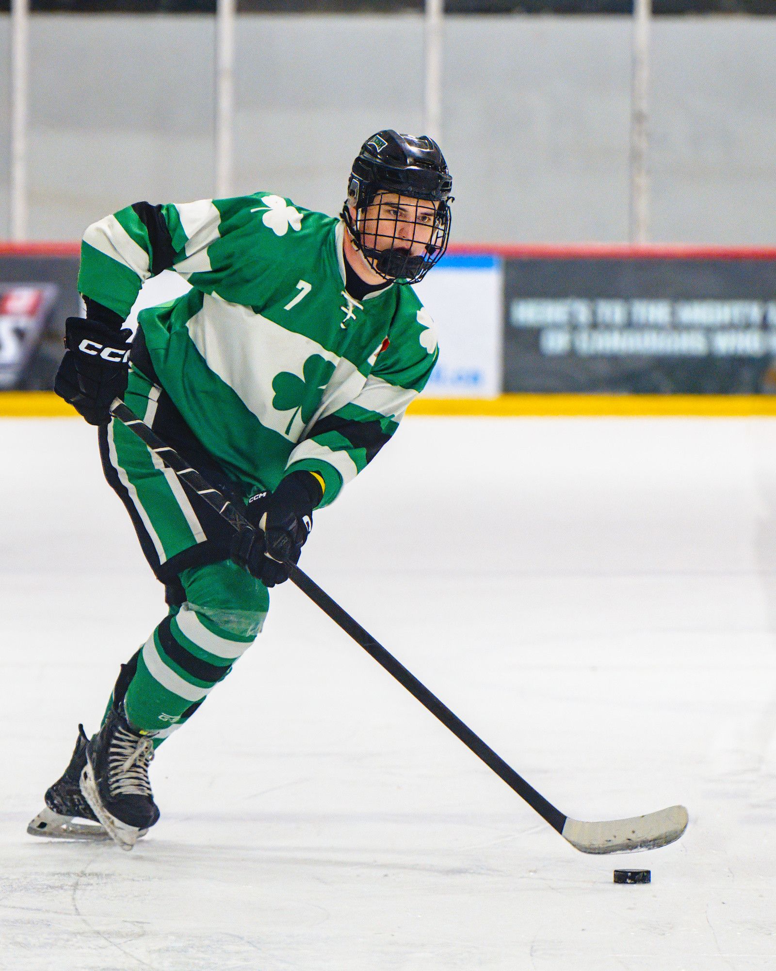 A forward in a green shamrock jersey carries the puck up the ice with his head up, reading the play in front of him.