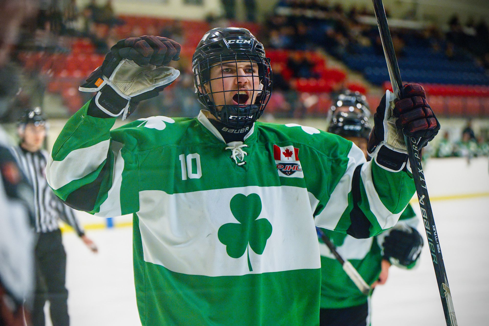 A player in a green shamrock jersey roars with one glove raised and his stick lifted, red arena seats behind him.