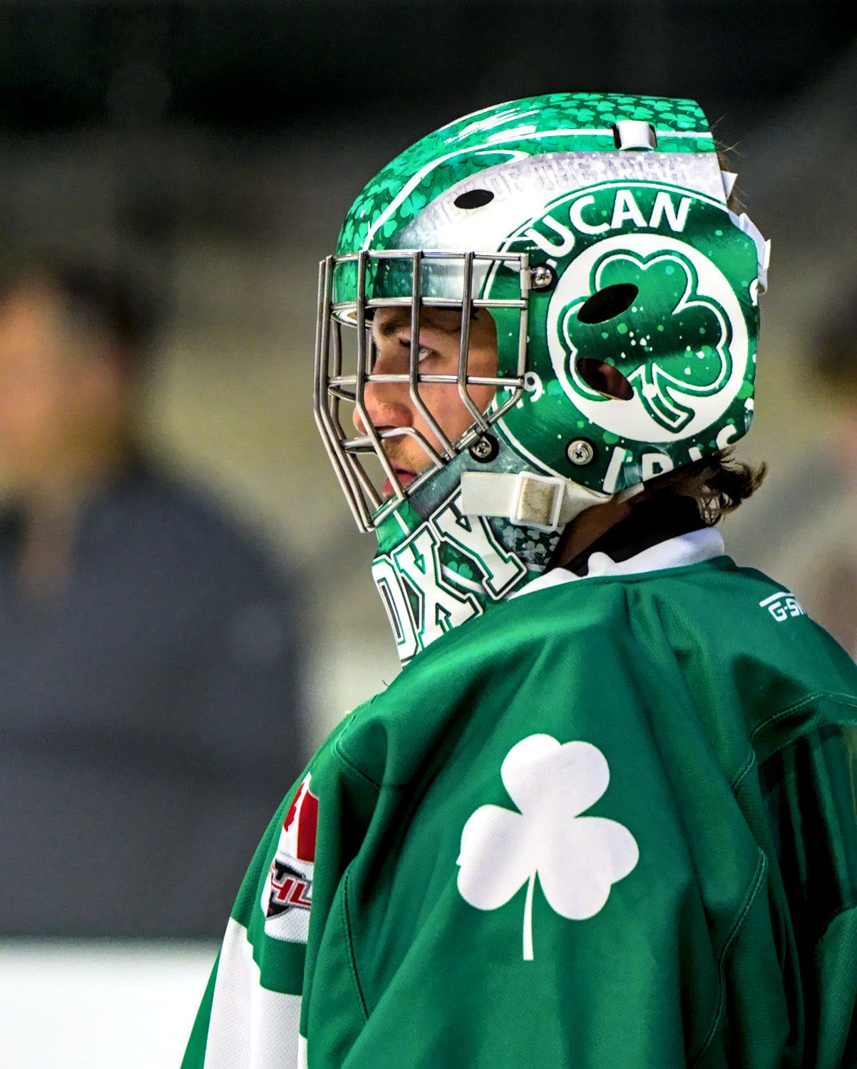 A goaltender in profile wearing a hand-painted green shamrock mask, eyes turned down the ice.