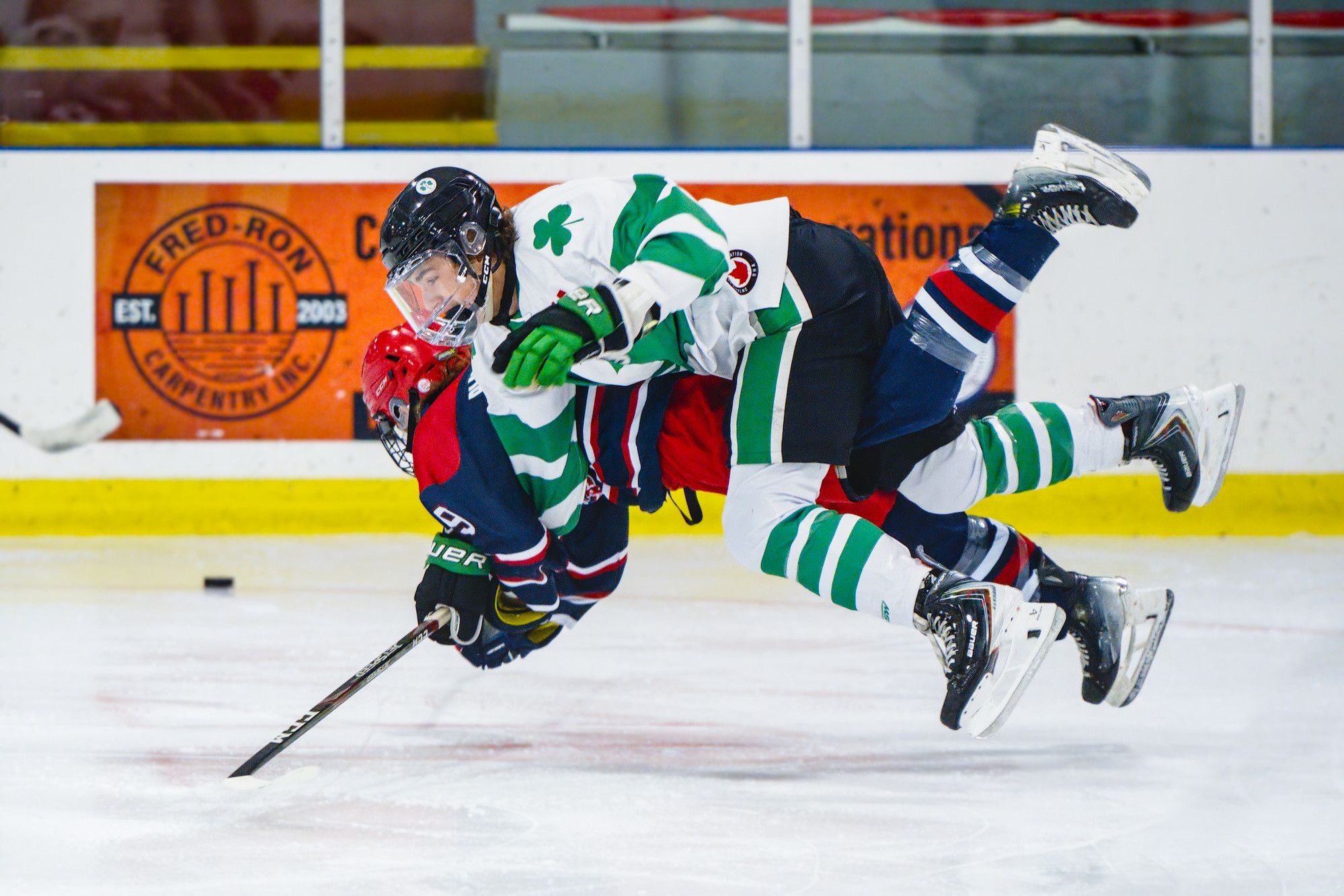 An open-ice hit lifts a player in a navy and red jersey off his skates as a shamrock-jersey forward drives through the check.