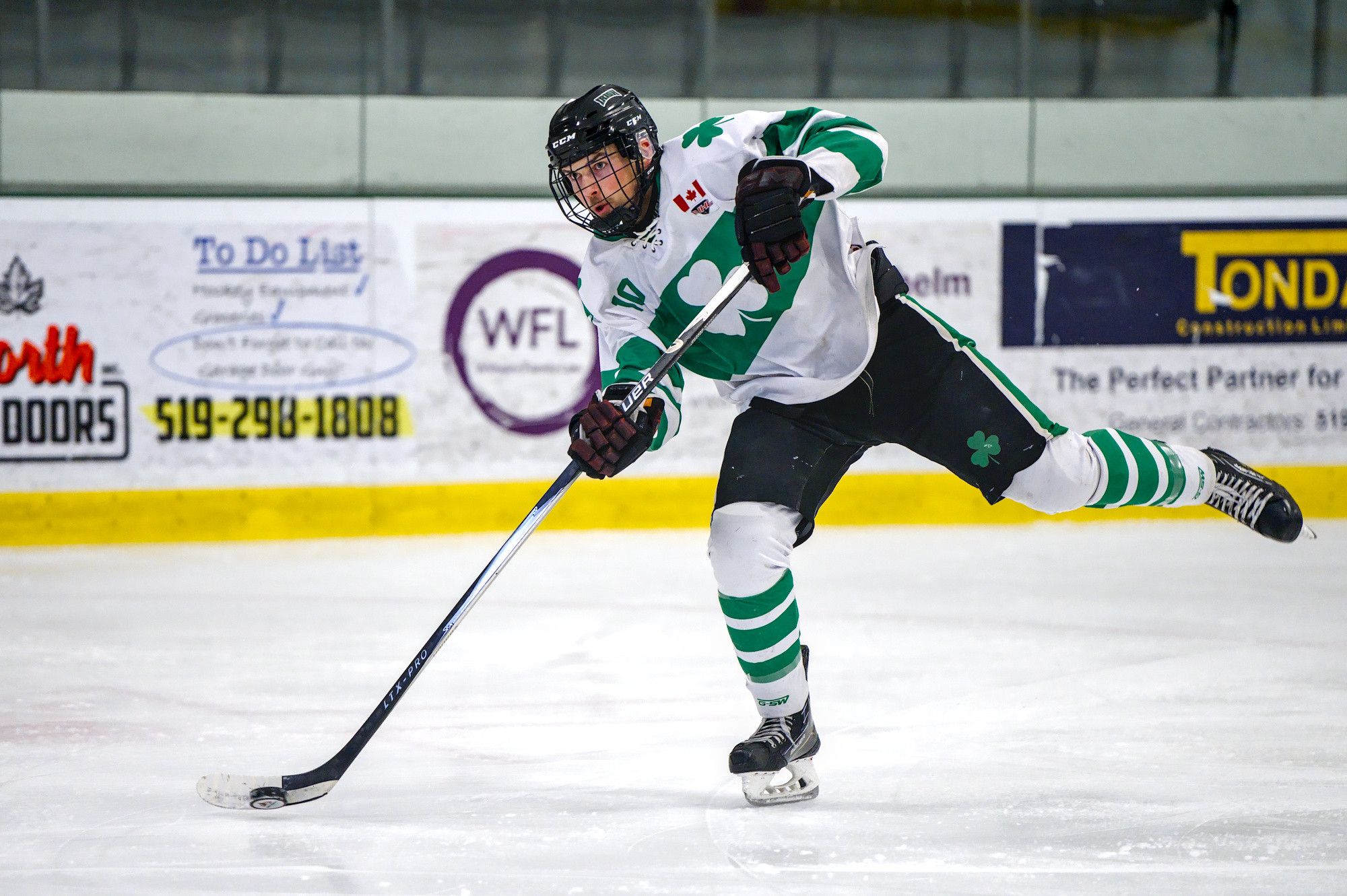 A shooter in a white shamrock jersey finishes a slapshot, back skate off the ice and the stick still coming through.