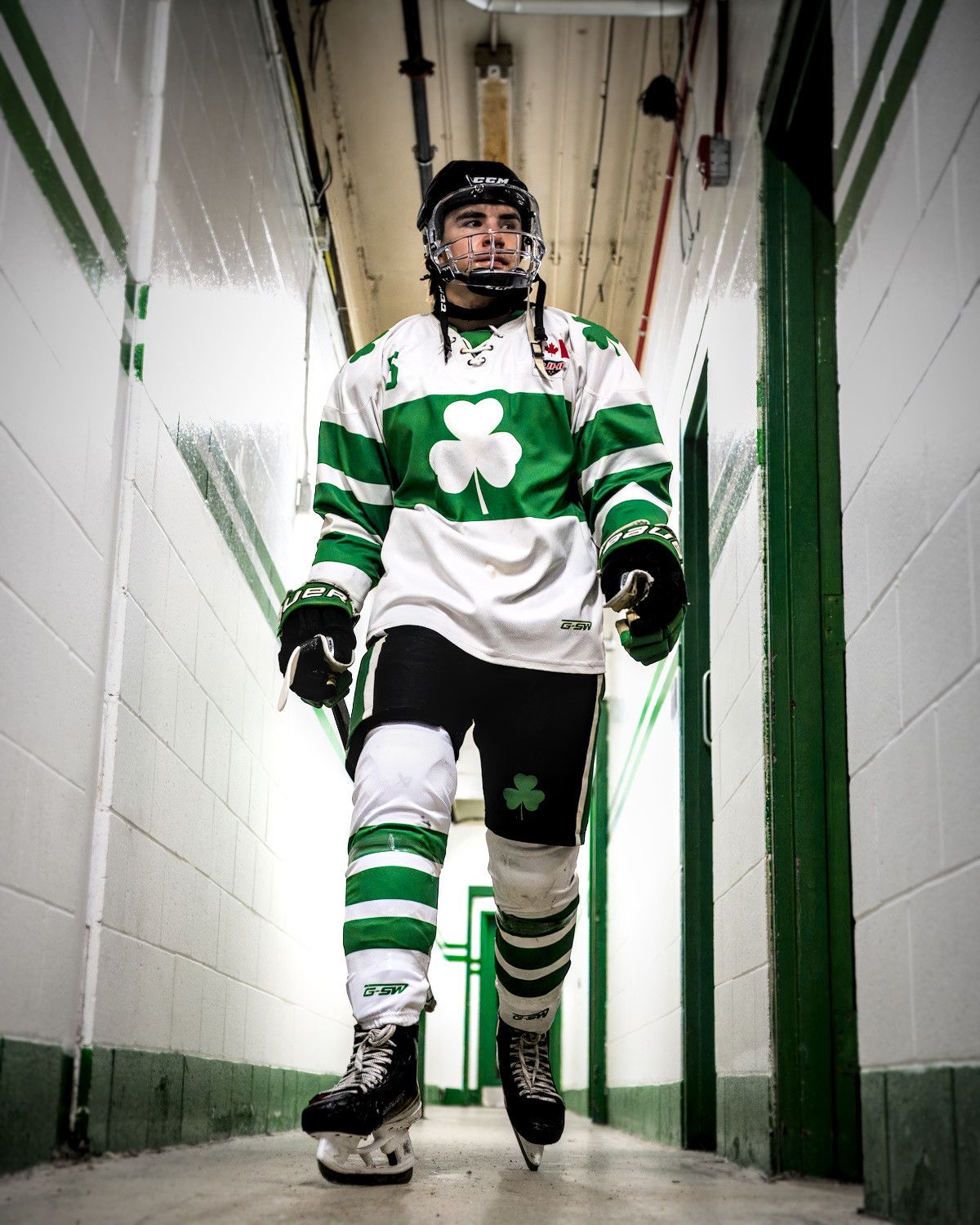 A player walks up the arena tunnel toward the ice in full gear, helmet on, gloves on and stick in hand, cinderblock walls close on both sides.