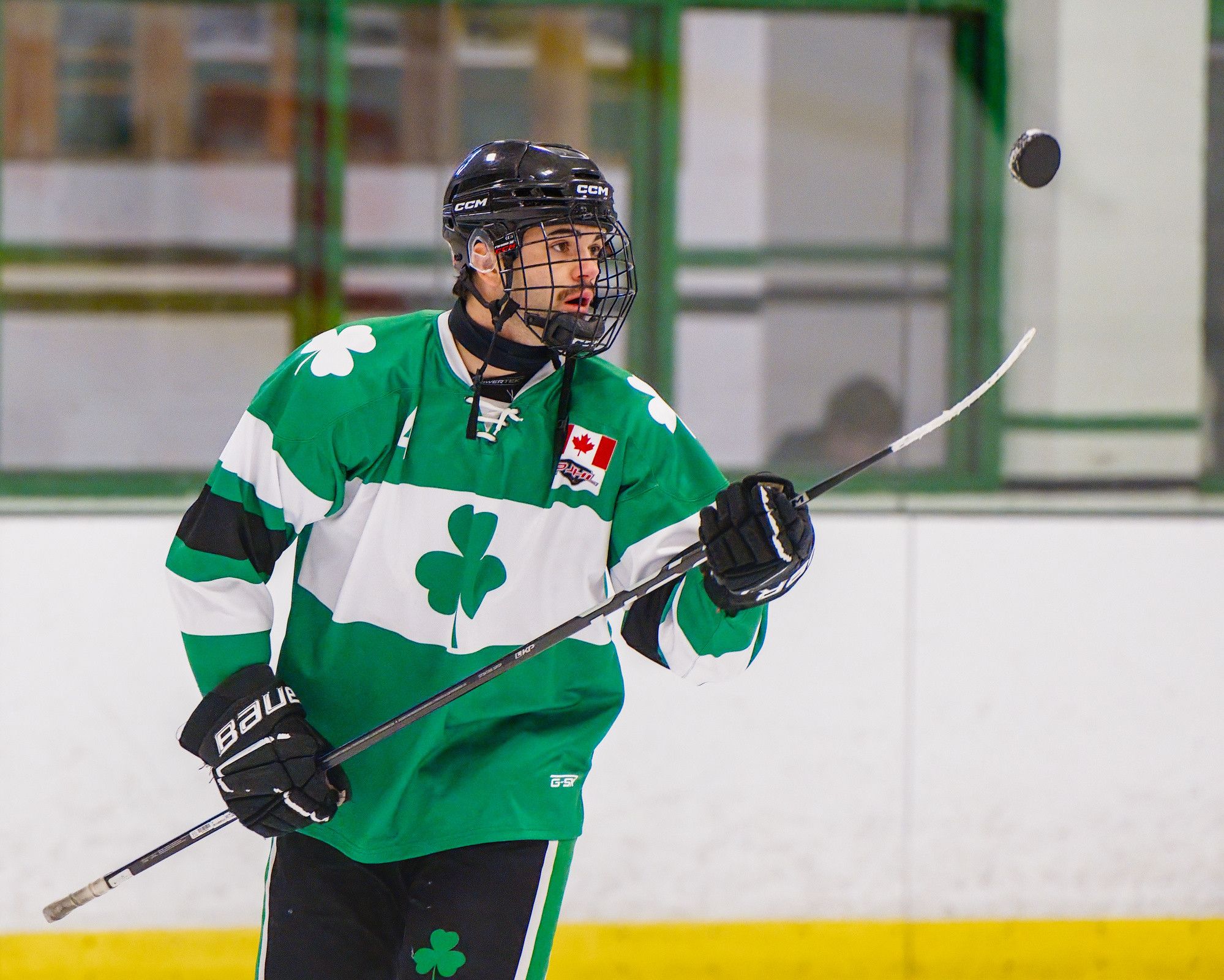 A moustached player in a green shamrock jersey tracks a puck he has flipped spinning in mid-air.